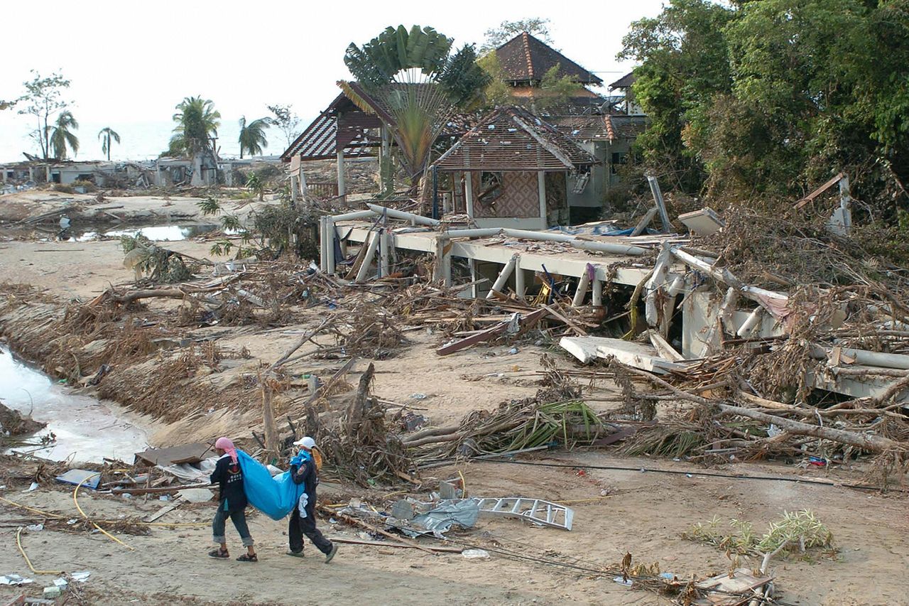 Two Thai rescuers carry a body recovered from the destroyed Khao Lak Laguna Resort Hotel, seen in the background, north of the devastated Thai tourist resort island of Phuket in December 2004Credit: ROMEO GACAD/AFP via Getty