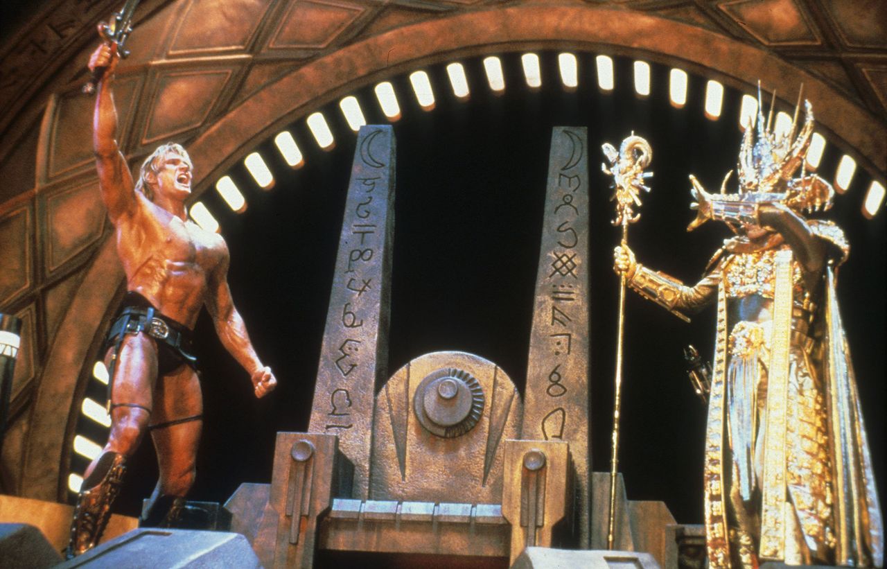 A man holding a sword and raising his fist confronts a robed figure carrying a staff on a thronelike set Dolph Lundgren as He-ManCredit: Everett