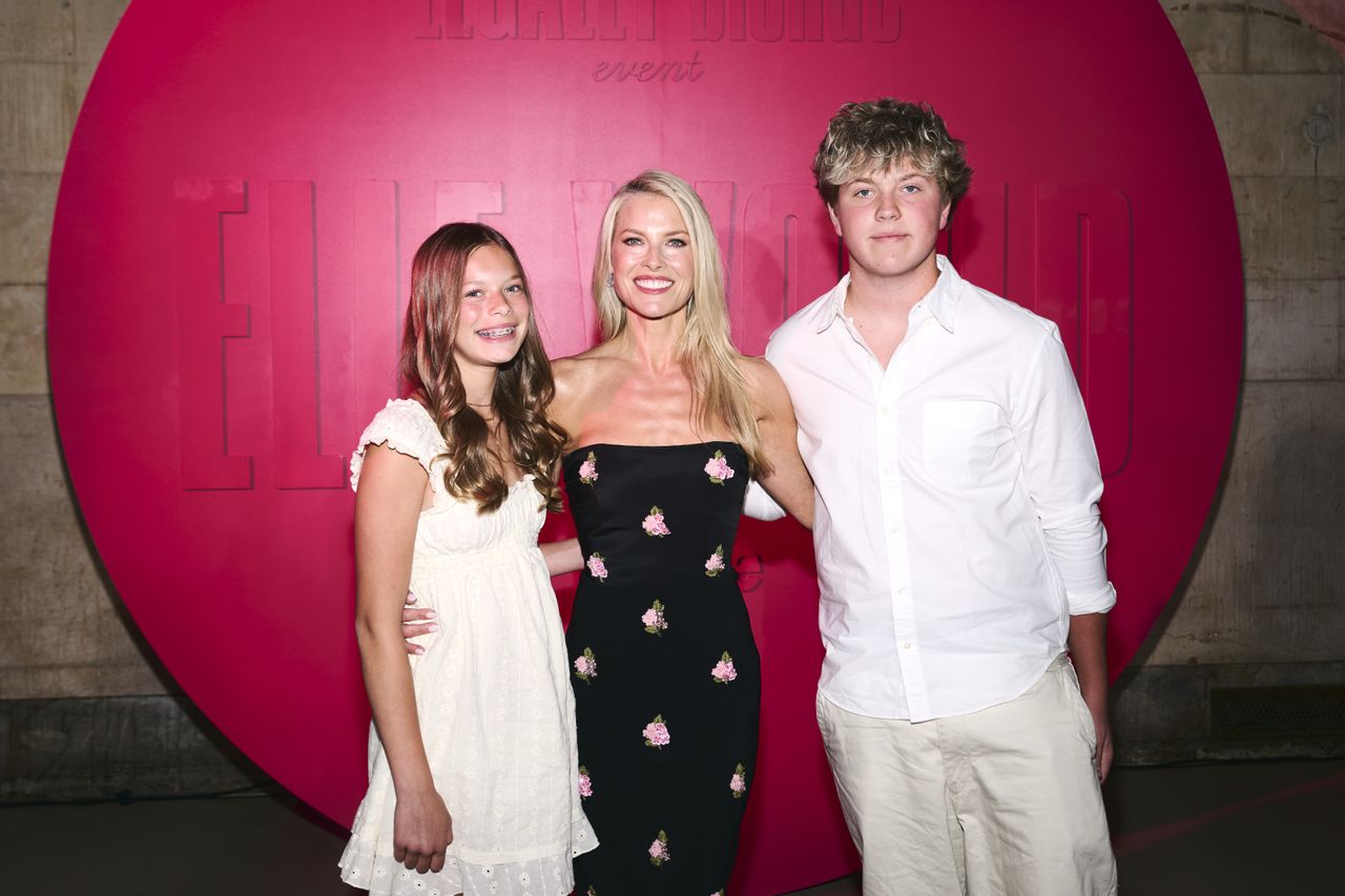 Ali Larter stands with her children on either side in front of a hot pink background. Larter's daughter, Vivienne, is on her right in a white ruffled minidress, while her son, Teddy, is on her left in a white collared shirt and khaki pants. Larter is wearing a strapless black midi dress embellished with pink sequin flowers. Ali Larter and her kids, Theodore