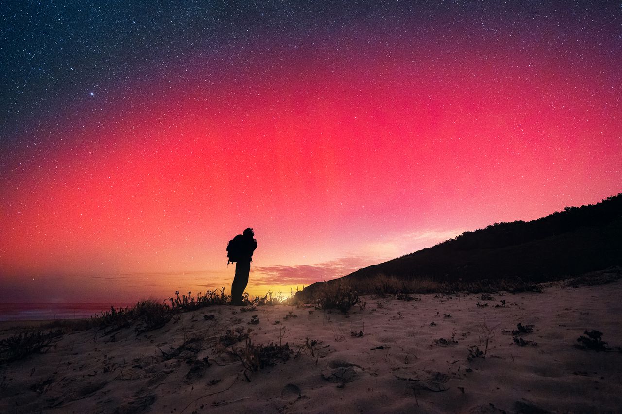 A backpacker stands on a sandy dune in Galicia, looking up at the northern lights. The night sky is illuminated with shades of red, pink, and orange as the lights dance above him. It's a rare sight in these latitudes, as auroras are uncommon in this region. Northern lightsCredit: Getty