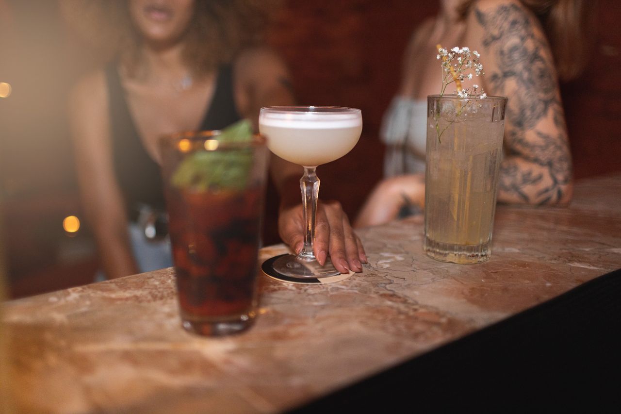 Three cocktails on a bar counter two women partially visible in the background engaging Women at a bar (stock image).